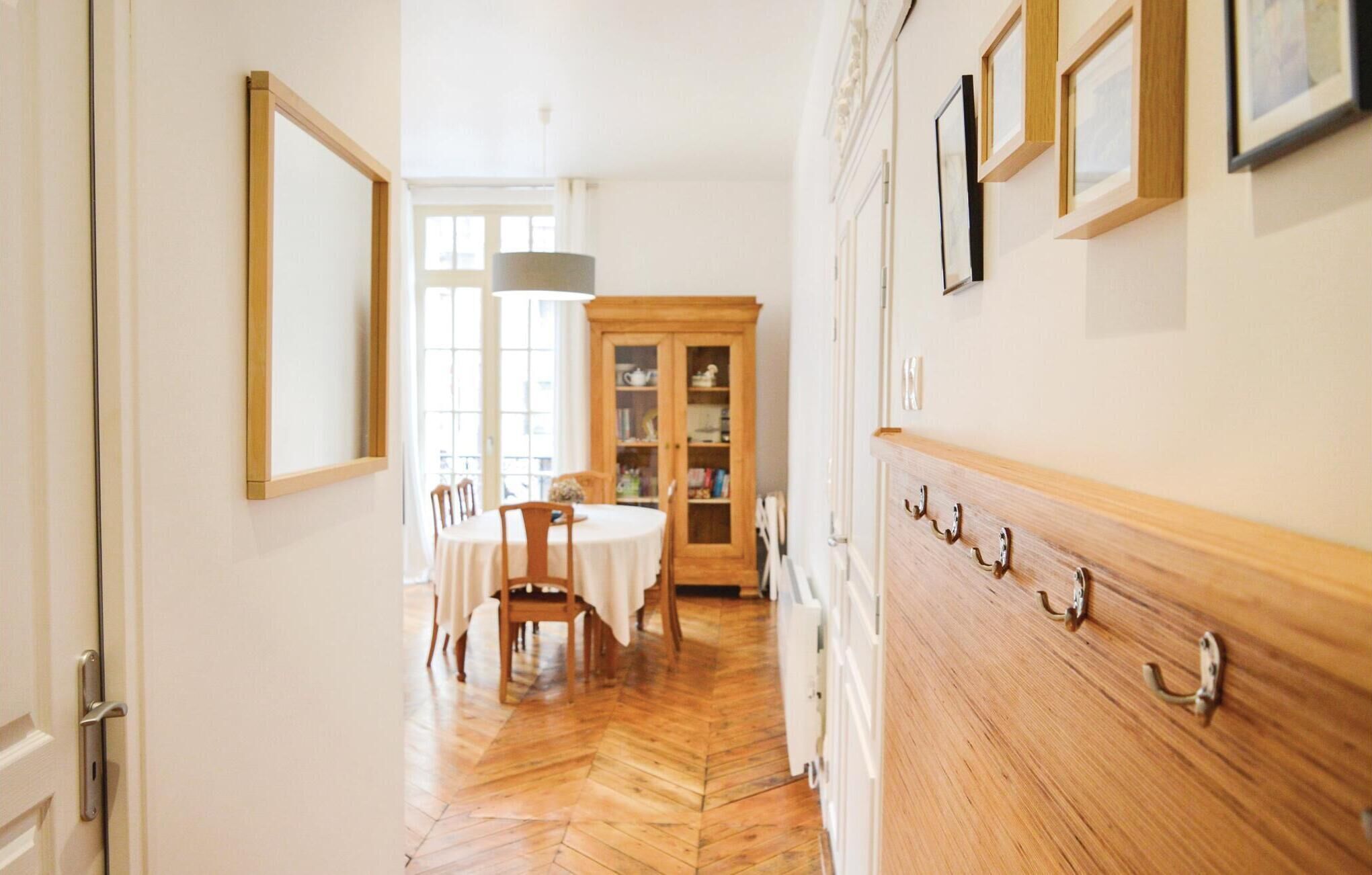 A hallway with a wooden floor, a dining area with a table and chairs, a cabinet with glass doors, and a wall with hooks.
