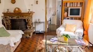 Un salon avec une cheminée en pierre, un canapé blanc, une table basse en verre sur laquelle sont posés des magazines, et un téléviseur sur un meuble en bois.