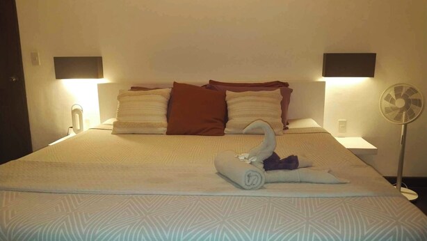 A neatly made bed with a swan towel decoration, two bedside lamps, and a standing fan. - Villa Kalachuchi B&B (Puerto Princesa)