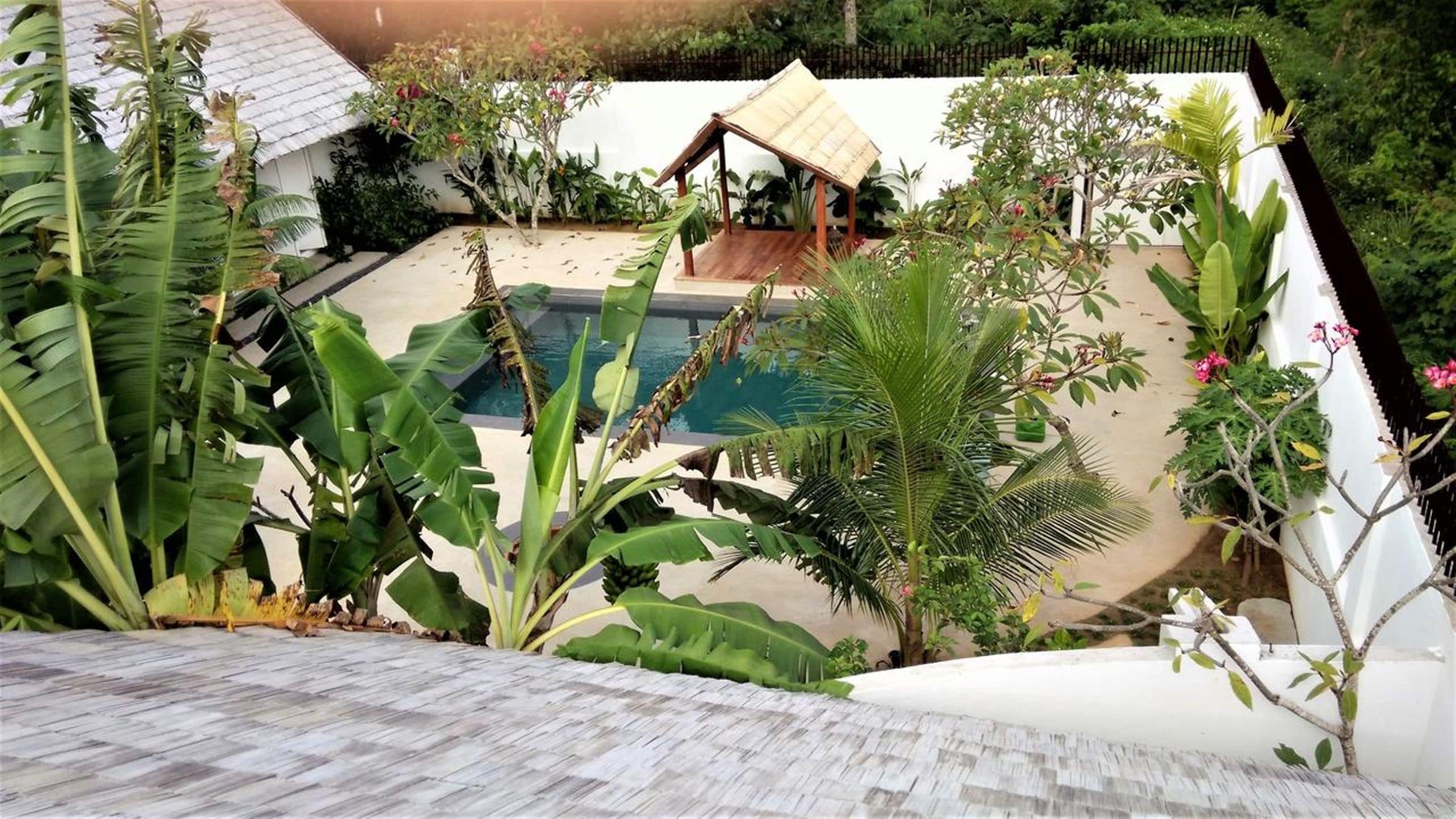Uma área de piscina com um deck de madeira, cercada por plantas tropicais e uma pequena construção de madeira.