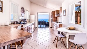 A modern kitchen with a large wooden table, white chairs, and a view of the sea through an open door.