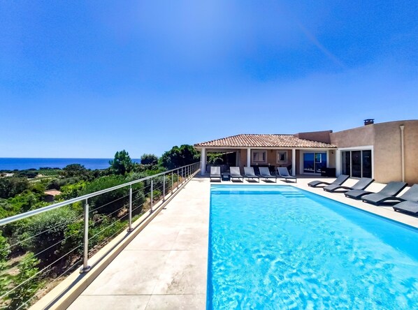 Une piscine avec des chaises longues, un espace de détente couvert et une vue sur la mer.