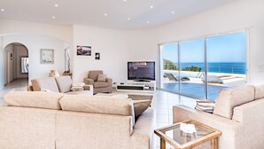 A modern living room with a large TV, comfortable seating, and a view of the ocean through floor-to-ceiling windows.