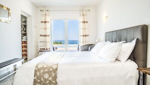 A bedroom with a large bed, a view of the sea through a sliding door, and a small seating area with a chair and table.