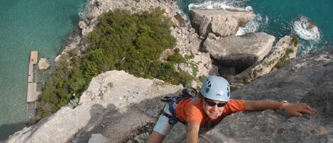 A person climbing a rocky cliff with a rope, overlooking a coastal area with clear water and greenery.
