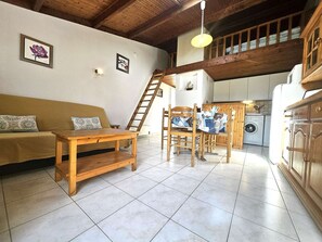 A cozy living area with a sofa, wooden table, and chairs, a kitchen with a washing machine, and a loft bedroom with a wooden ceiling.