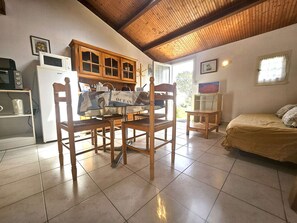 A dining area with wooden chairs and a table, a kitchen with appliances, a bed with a beige cover, and a TV on a stand.