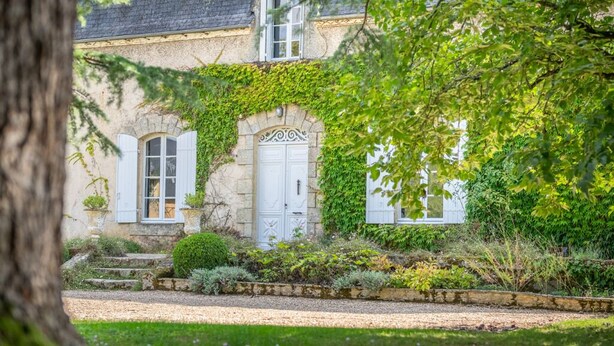A stone building with white shutters and a central door, surrounded by lush greenery and flowering plants.