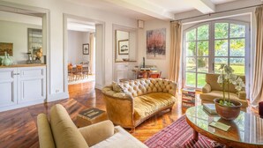 A spacious living room with a large window, a sofa, a coffee table, and a dining area visible in the background.
