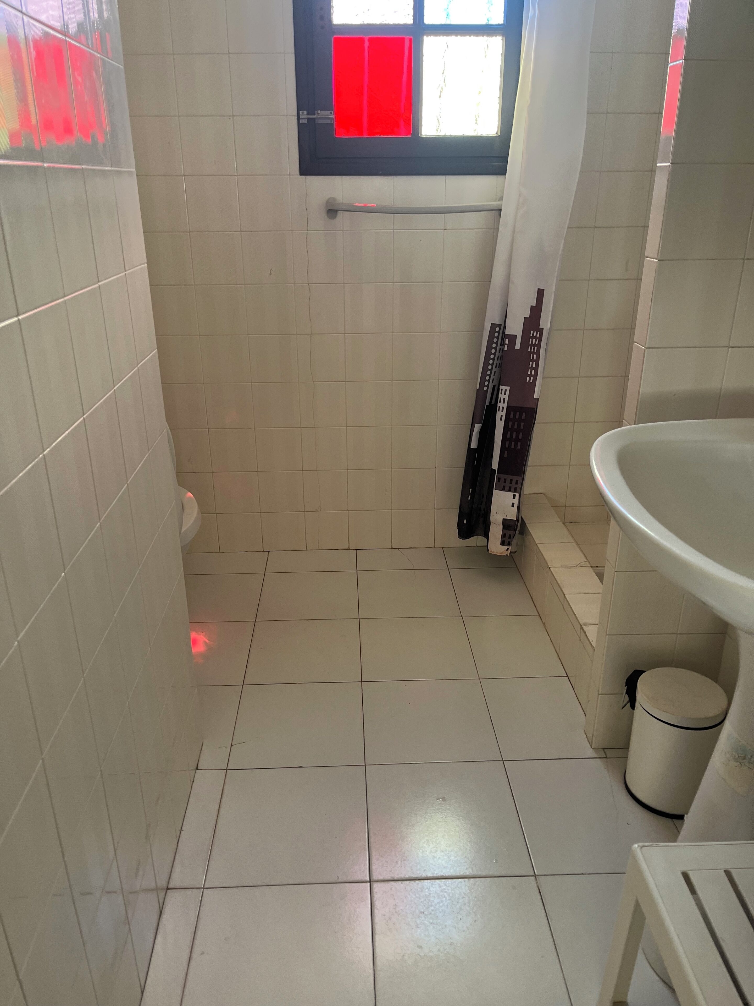 A bathroom with white tiled walls, a window with red curtains, a toilet, and a sink.