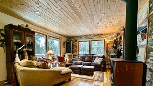 A cozy living room with a fireplace, wooden floors, and large windows.