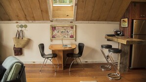 Acogedor dormitorio en el ático con techo de madera, una mesa de comedor con sillas, una pequeña cocina con refrigerador y una ventana con vistas a árboles.