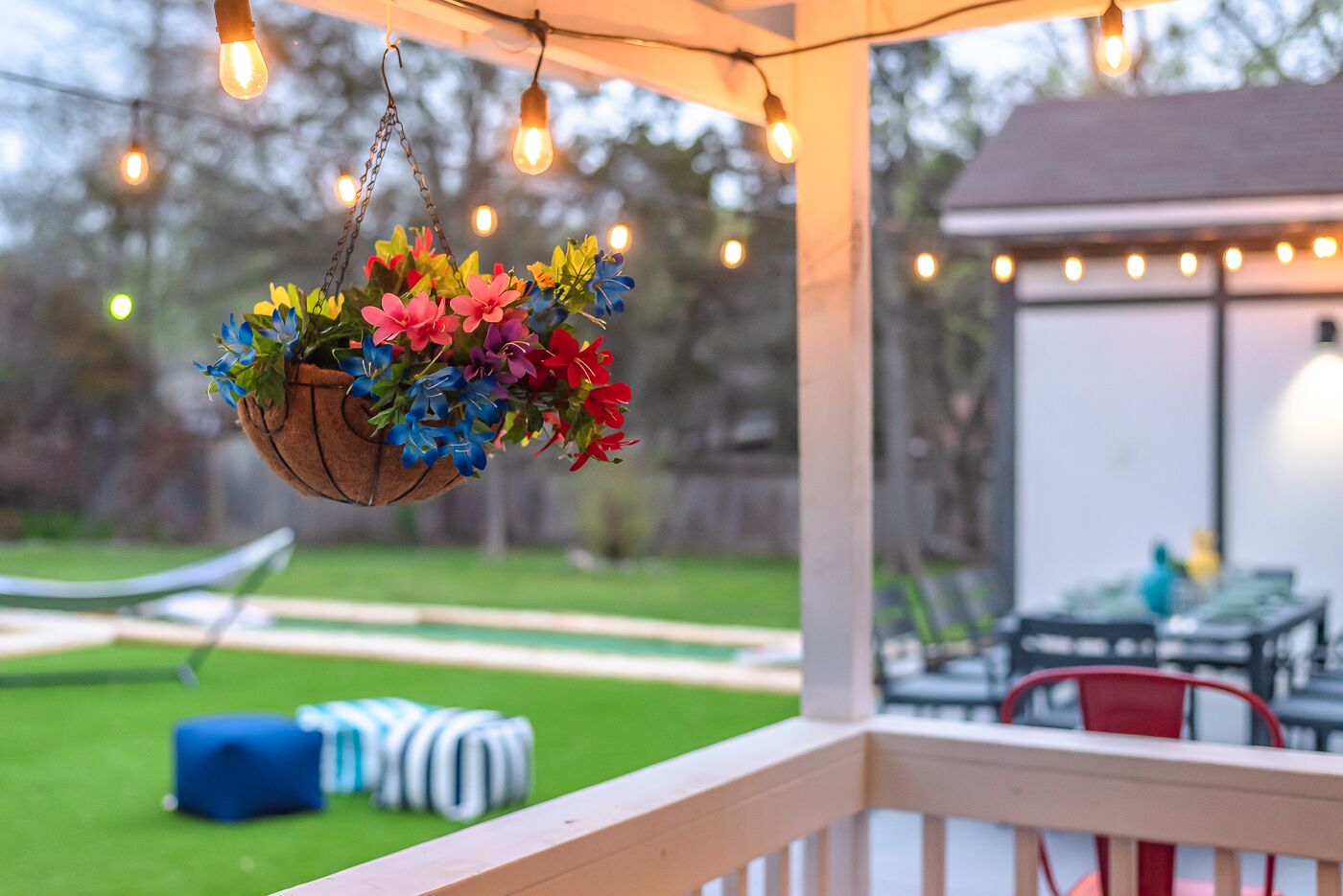 Una cesta colgante con diversas flores coloridas, una terraza de madera y luces cálidas al fondo.