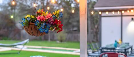 Una cesta colgante con diversas flores coloridas, una terraza de madera y luces cálidas al fondo.