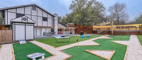 A modern house with a backyard featuring a putting green, a hammock, and a barbecue area.