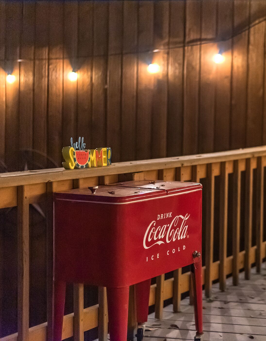 Un enfriador de hielo rojo con la marca Coca-Cola sobre una terraza de madera, con un cartel que dice 'Hello Summer'.