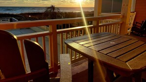 Um deck de madeira com mesa e cadeiras, com vista para uma praia onde o sol se põe no oceano.