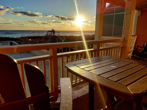 A wooden deck with a table and chairs, overlooking a beach with the sun setting over the ocean. - Bright and Beachy Oceanfront Condo - Linens included! (Oak Island)