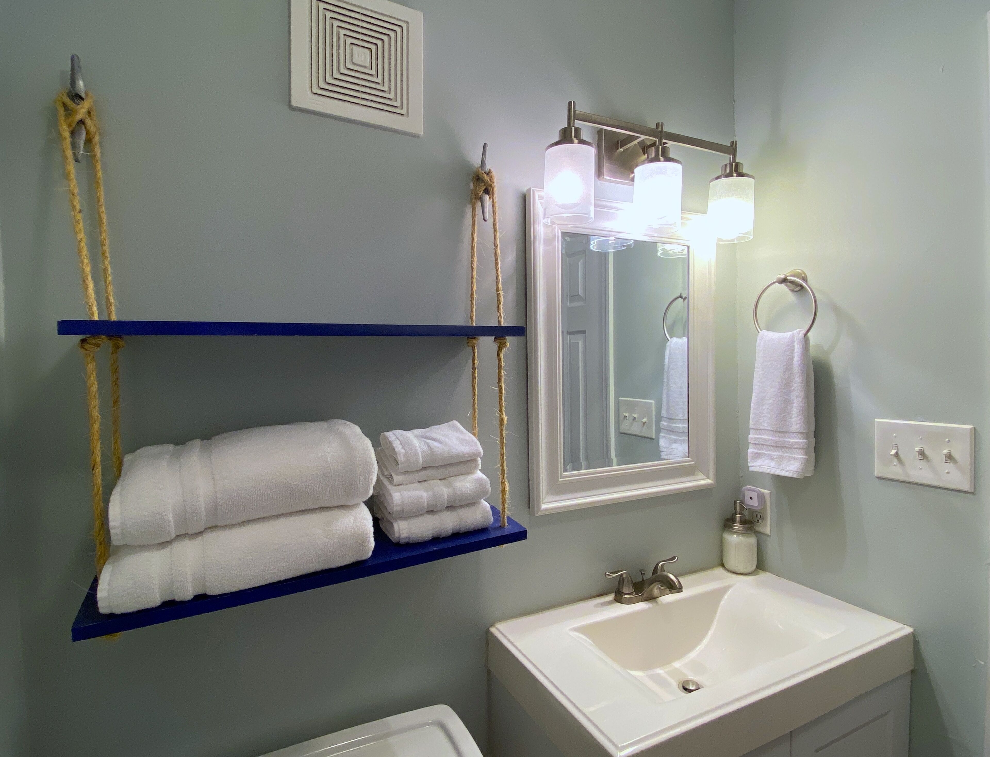 A bathroom with a white sink, a mirror with lights, and towels on blue shelves.