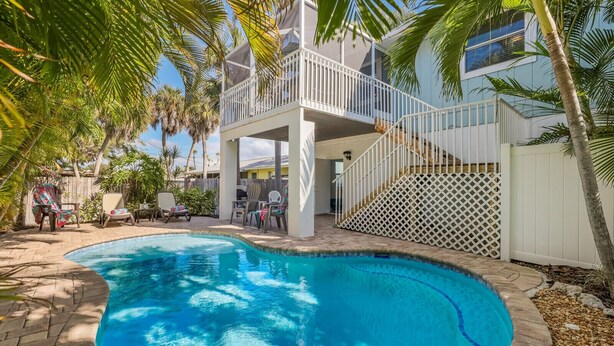 A swimming pool with a white deck, surrounded by palm trees and a wooden fence. - Island Hideaway East: 5 Minute Walk to Beach, Private Pool by Anna Maria Life Vacation Rentals (Holmes Beach)
