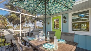 An outdoor patio with a table set for two, surrounded by wicker chairs, under a patterned umbrella. - Island Hideaway East: 5 Minute Walk to Beach, Private Pool by Anna Maria Life Vacation Rentals (Holmes Beach)