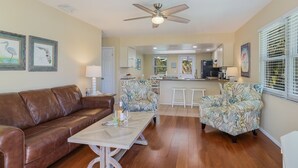 A living room with a brown leather couch, two floral armchairs, a wooden coffee table, and a ceiling fan. - Island Hideaway East: 5 Minute Walk to Beach, Private Pool by Anna Maria Life Vacation Rentals (Holmes Beach)