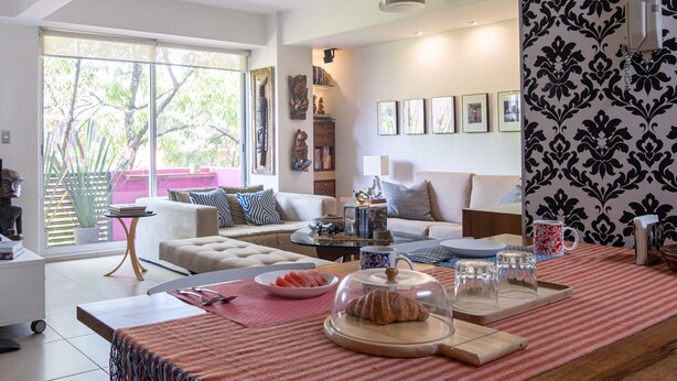 Una sala de estar moderna con un sofá, una mesa de centro y un comedor. Una ventana grande ofrece luz natural y vistas a árboles.