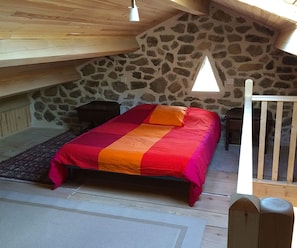 Un dormitorio con pared de piedra, techo de madera, cama con manta roja y naranja, y una ventana pequeña.
