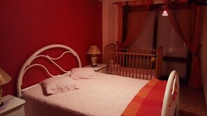 A bedroom with a single bed, a nightstand, a lamp, a crib, and a window with curtains.