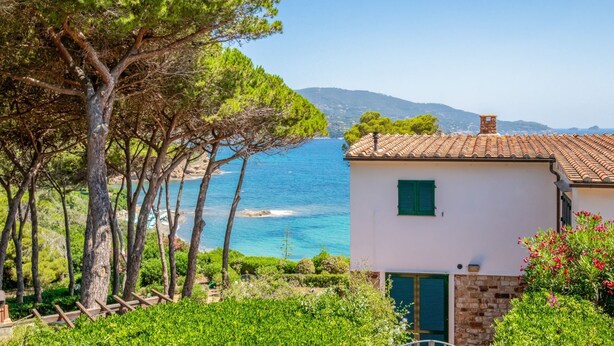 Une vue sur la côte, avec un bâtiment blanc aux volets verts, entouré d’arbres et bordé par une mer bleue et limpide.