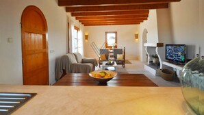Uma sala de estar acolhedora com teto de madeira, uma mesa de jantar com uma cesta de frutas, um sofá, uma TV e uma lareira.