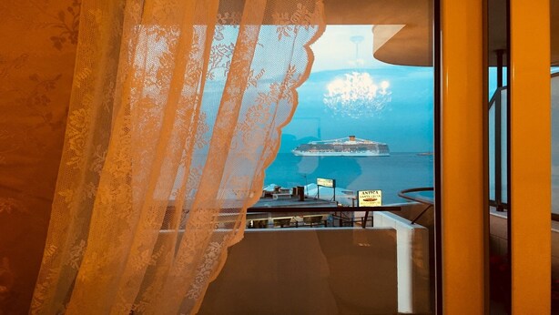 A balcony with a view of a cruise ship in the distance, a sign for 'ANTICA Santa Lucia Restaurante', and a lace curtain partially covering the view.