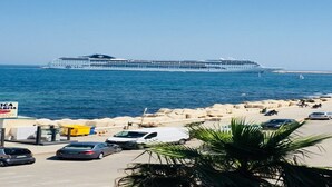 A cruise ship docked at a port with a rocky shoreline and parked cars.