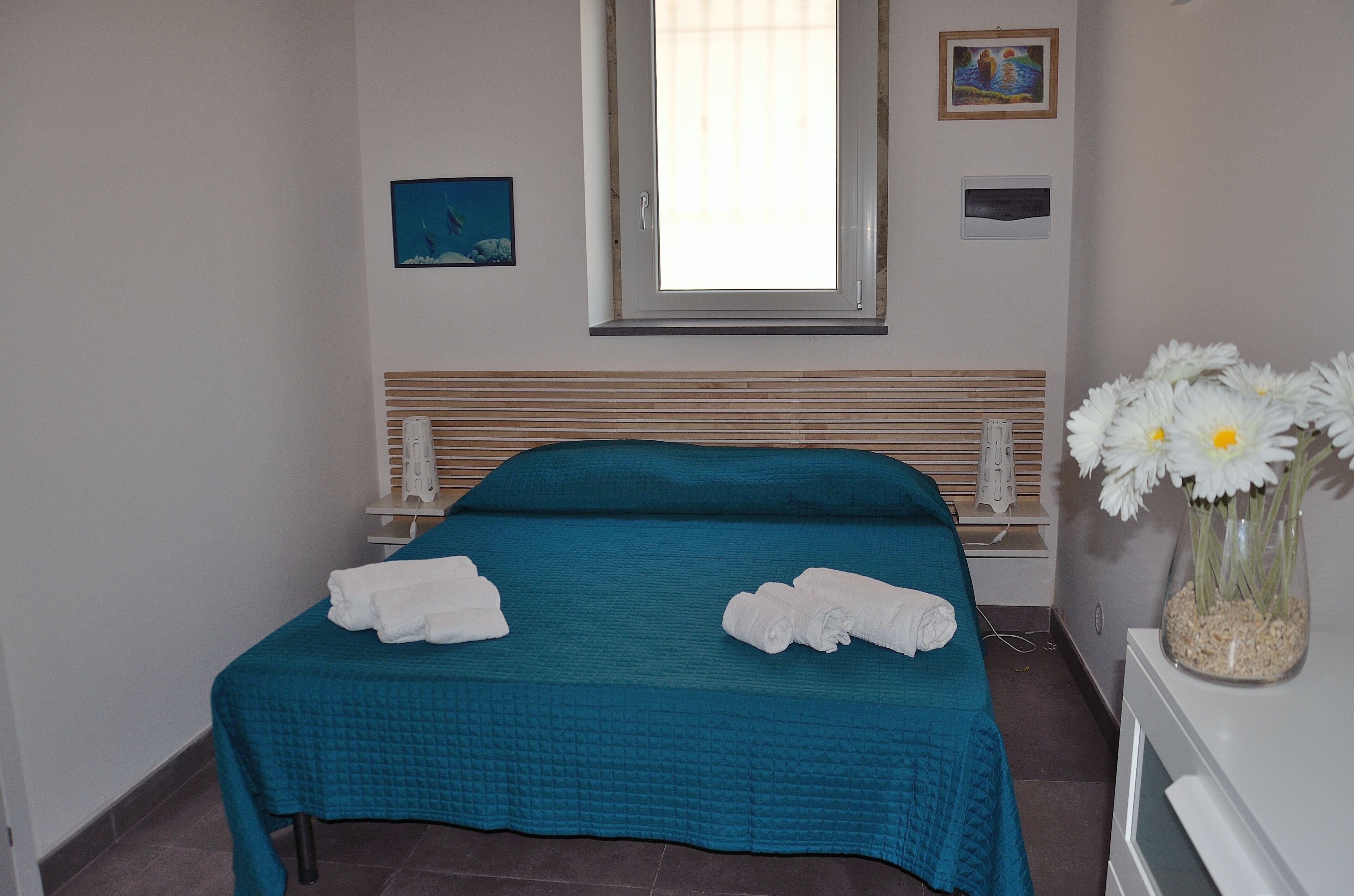 A neatly made bed with a teal bedspread and two rolled towels, a window with blinds, a small framed picture, and a vase with white flowers on a bedside table.