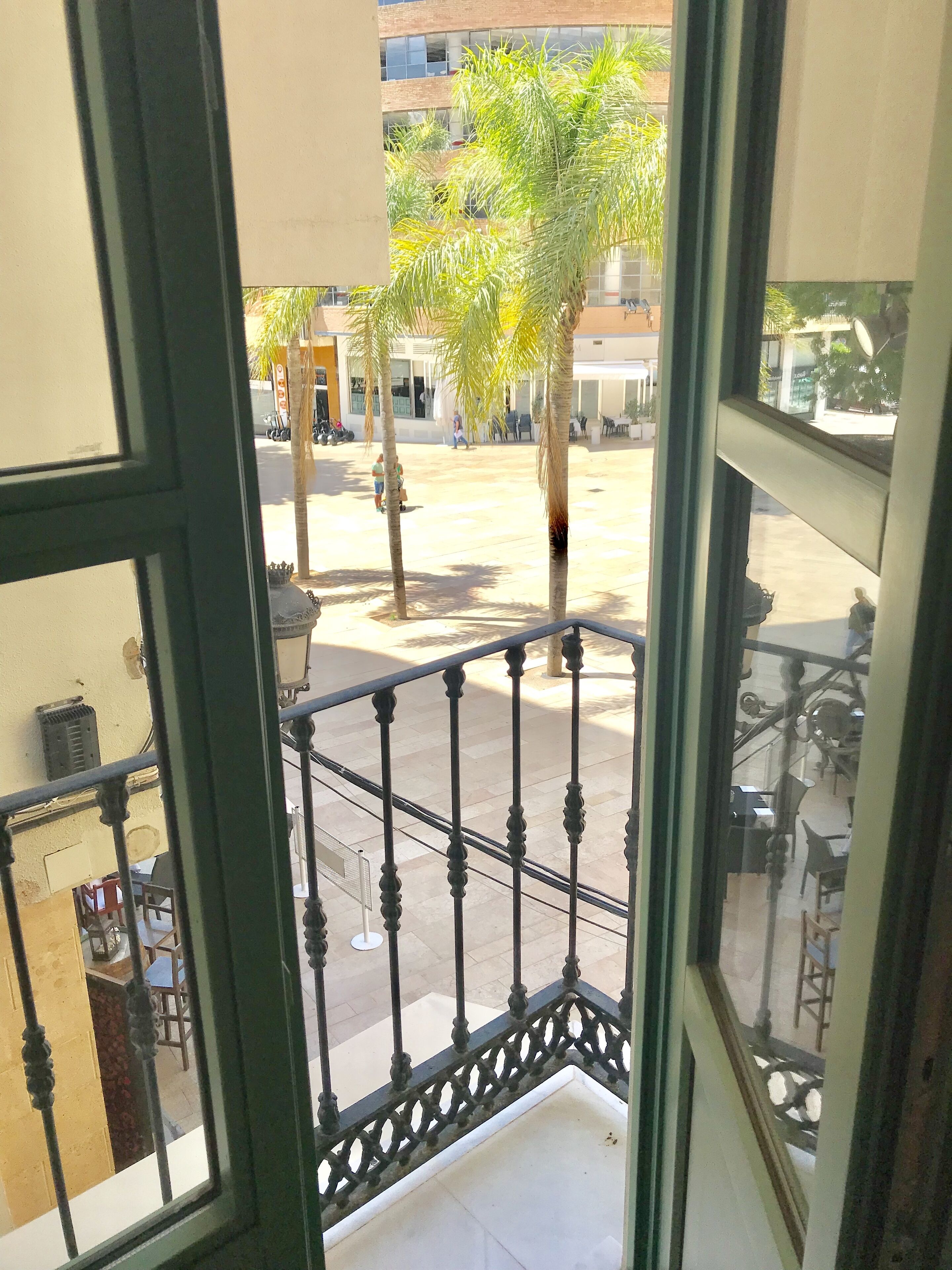 Vista desde un balcón con barandilla de hierro forjado, que da a un patio con palmeras y un edificio al fondo.