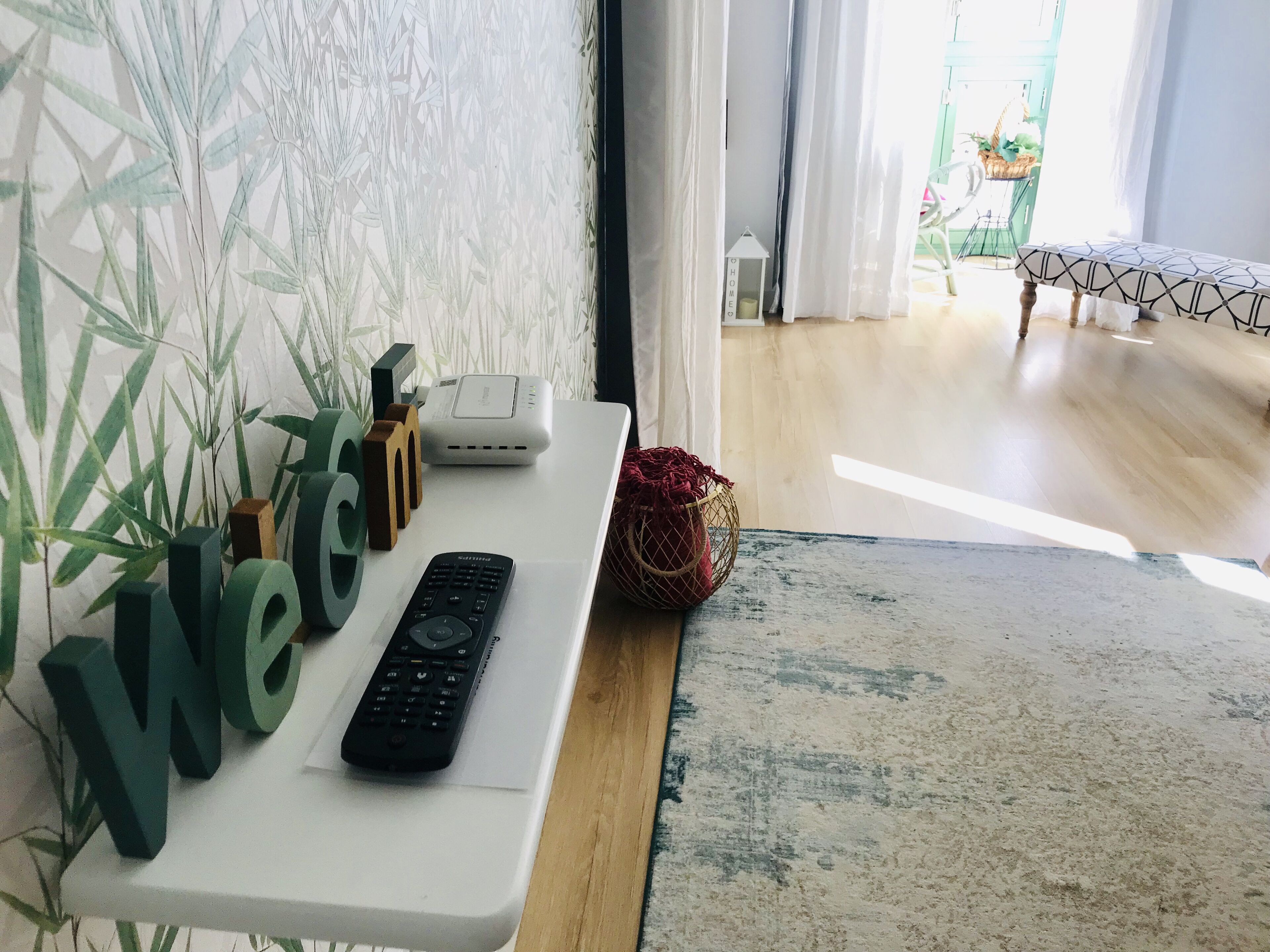 A room with a white console table holding a remote control, a decorative wall with large letters, a white device, and a basket.