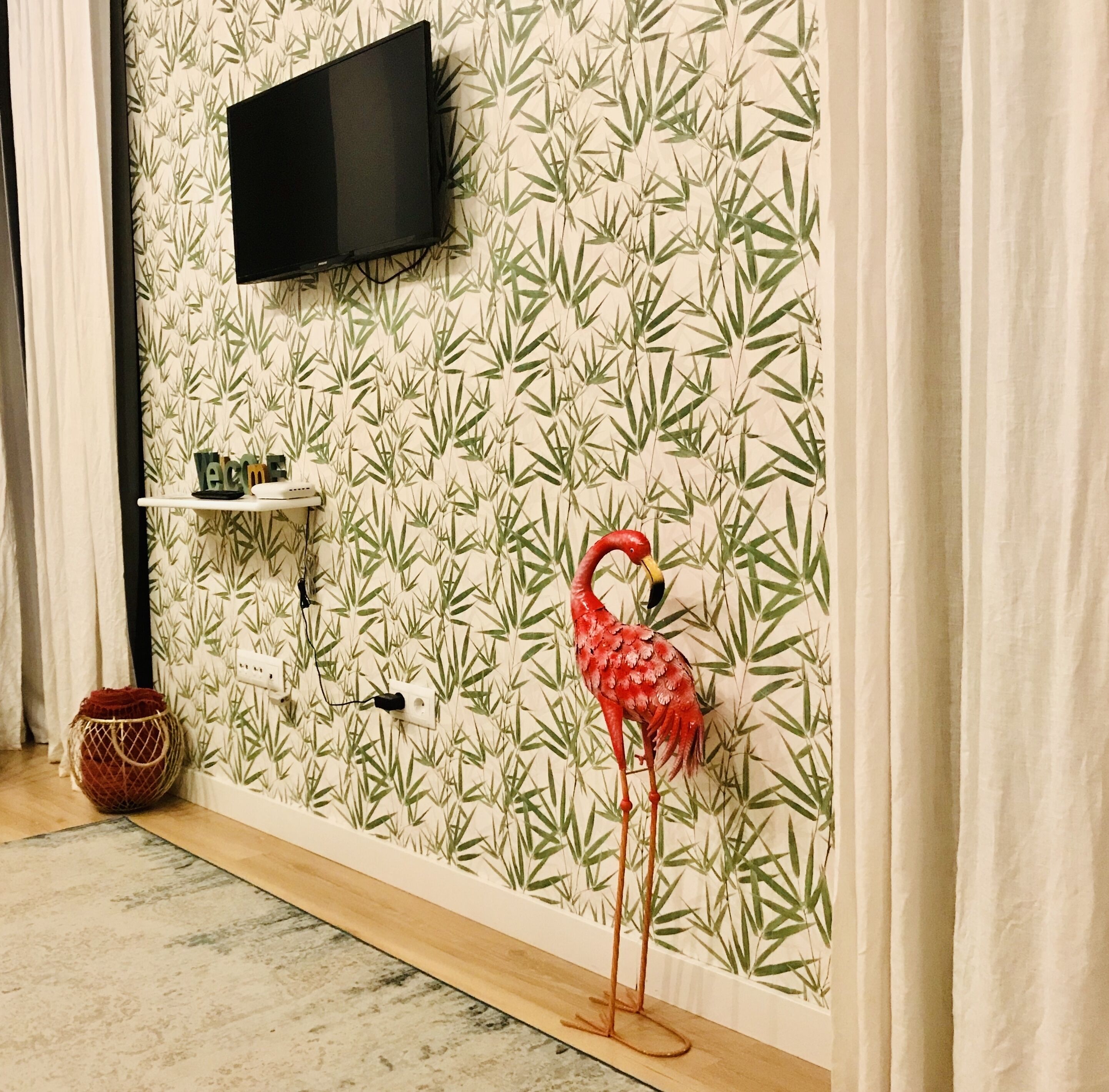 Ein Zimmer mit Bambus-Tapete, einem Wand-Fernseher, einer kleinen Ablage mit Dekoration und einer roten Flamingofigur.