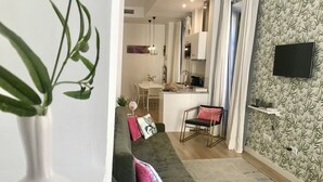 A modern living room with a green sofa, a small table with a bowl of white flowers, a kitchen area with a white countertop, and a TV mounted on the wall. -  Light and tranquility in the best location. Destiny Cathedral  (Málaga)