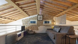 Une pièce mansardée chaleureuse avec un mur en pierre, des poutres en bois, un canapé, un fauteuil poire et un téléviseur.