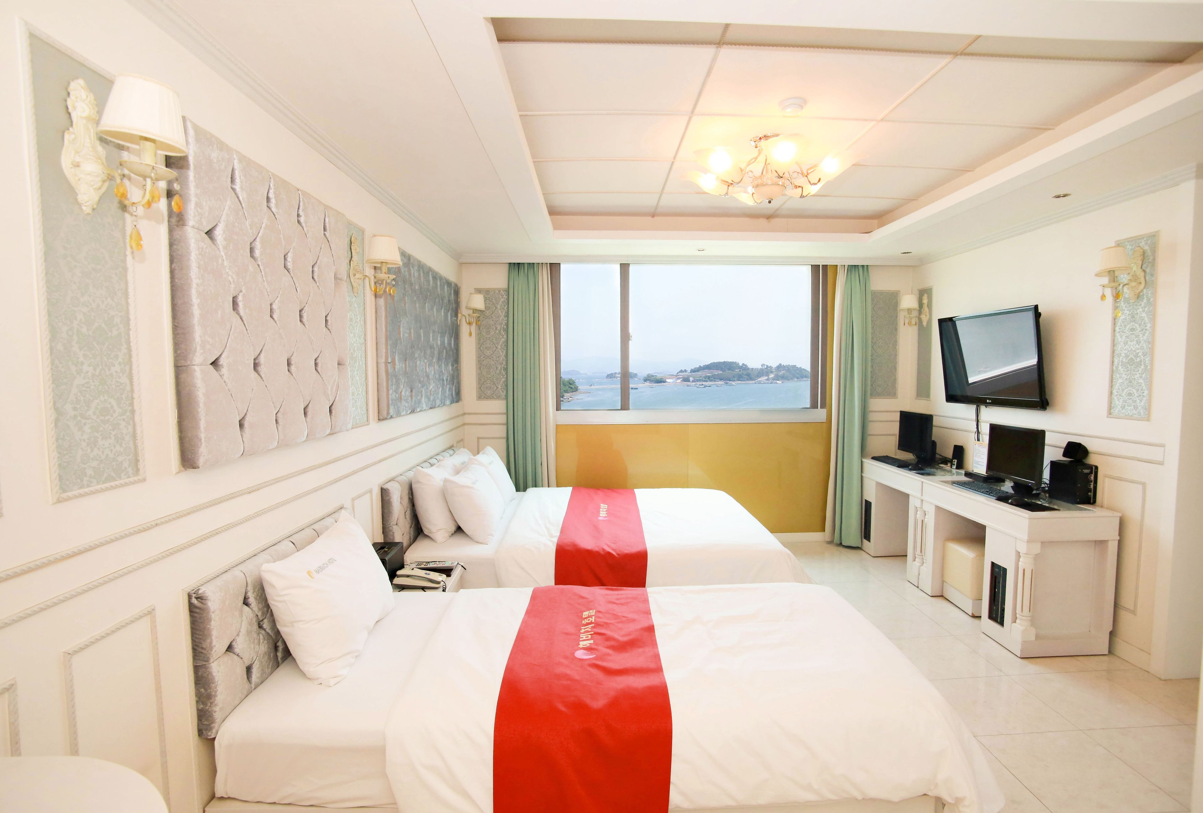 A hotel room with two beds, a desk with a TV, and a large window with a view of the sea.