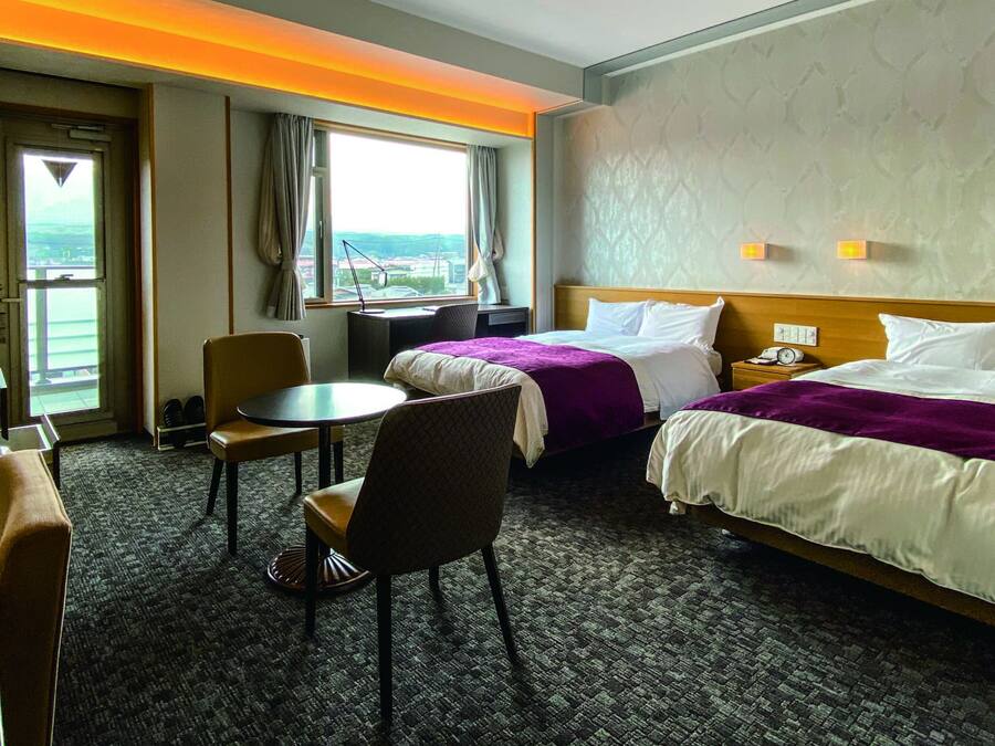 A hotel room with two beds, a desk, a chair, a small table, and a window with a view.