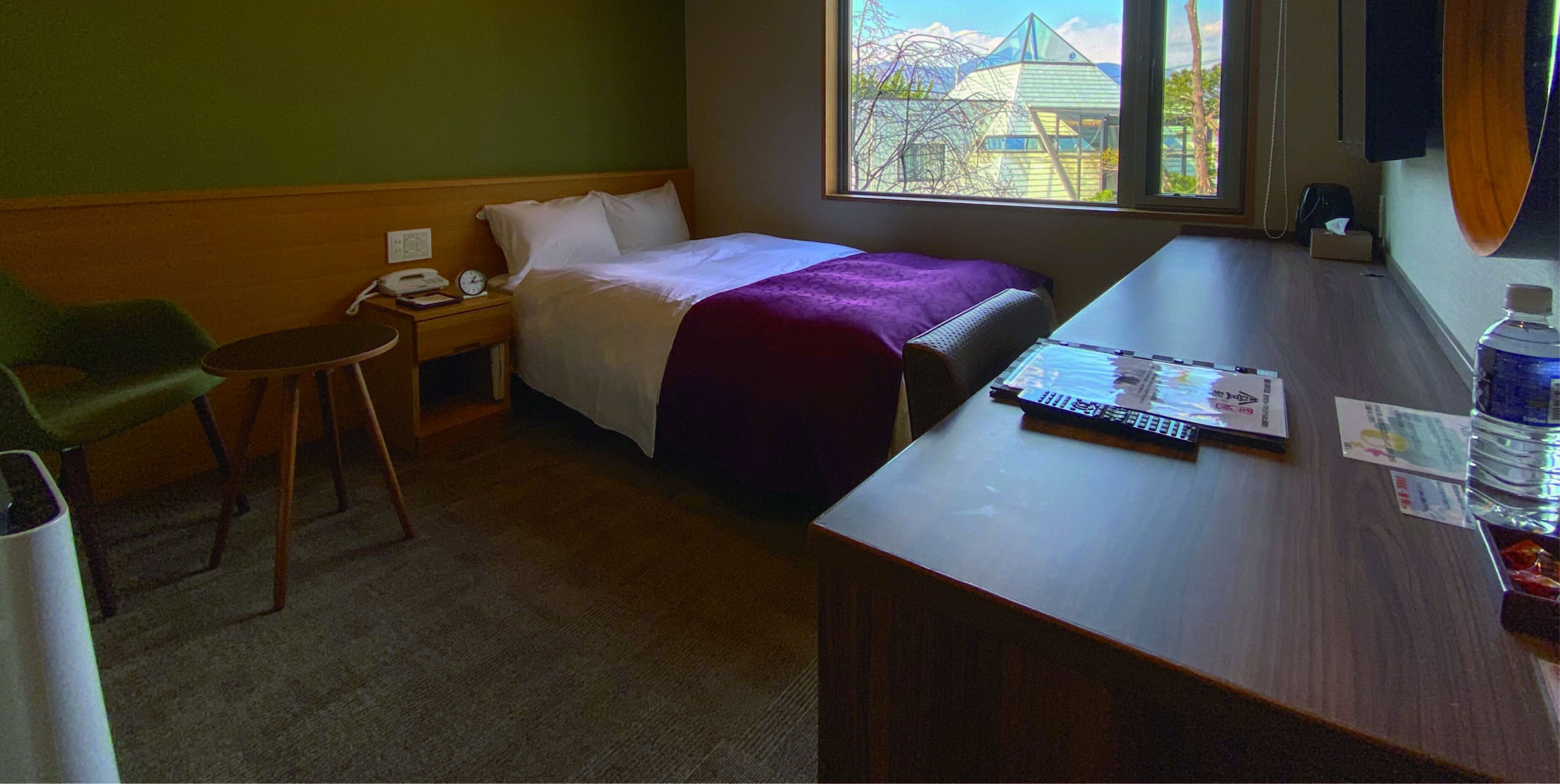 A hotel room with a bed, a desk, a chair, a nightstand, and a window with a view of buildings.
