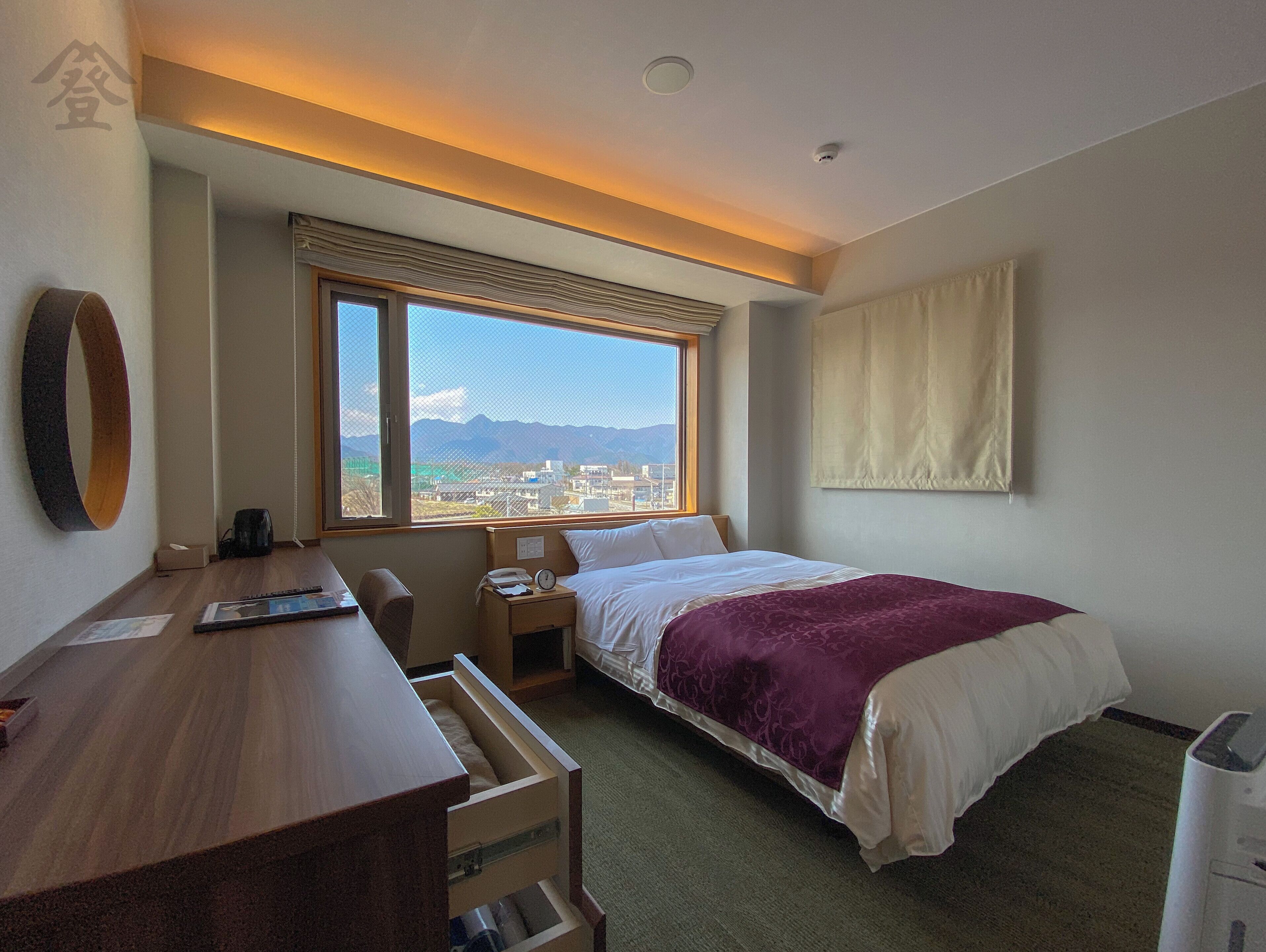A hotel room with a bed, a desk, a chair, a window with a view of a cityscape and mountains, and a ceiling light.