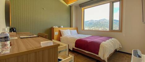 A hotel room with a bed, a desk with a chair, a window with a mountain view, and a wooden desk with a bottle of water and a notepad.