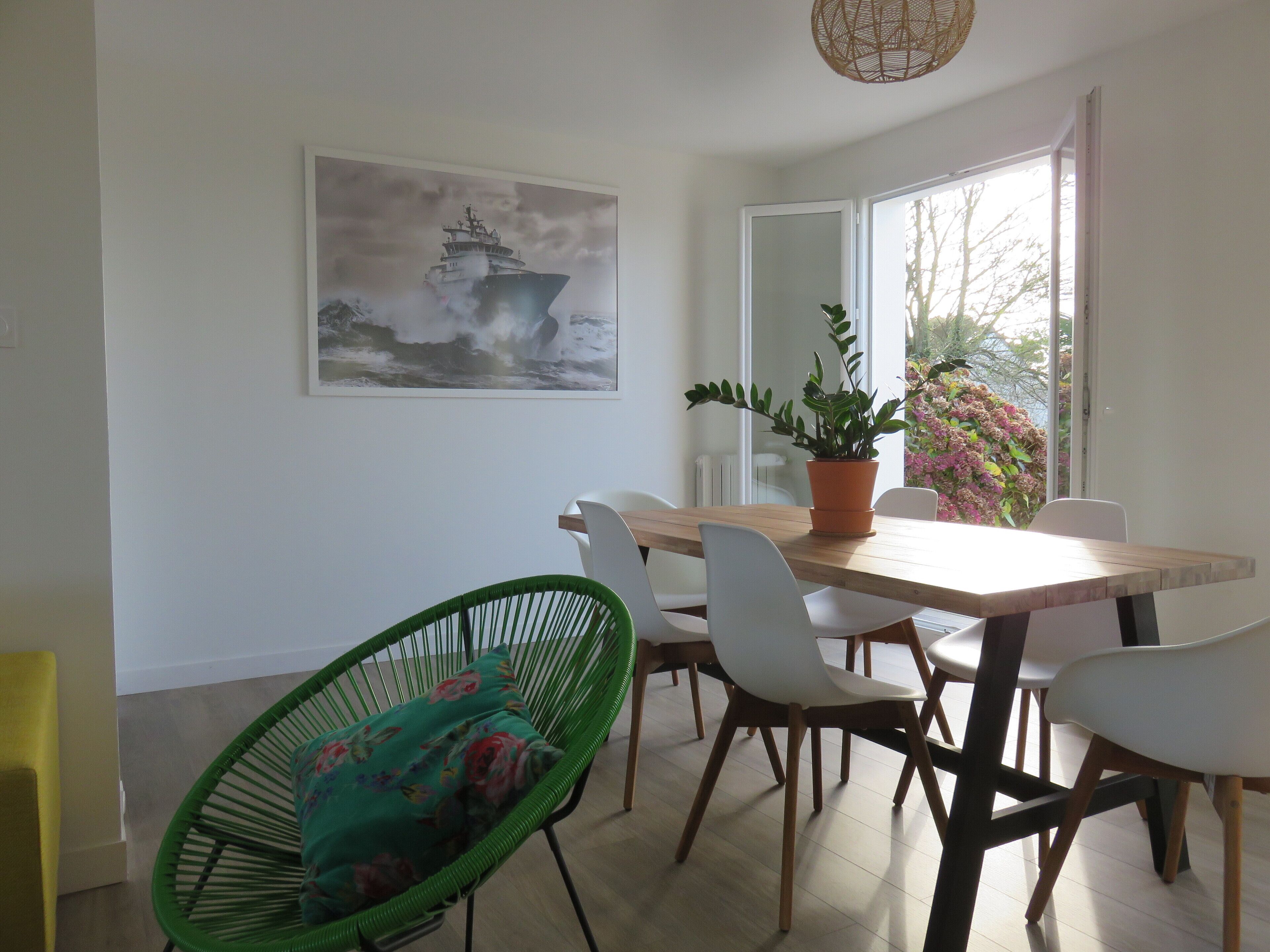 Un coin repas avec une table en bois, des chaises blanches, une chaise en osier verte, une plante en pot et un grand tableau représentant un navire accroché au mur.