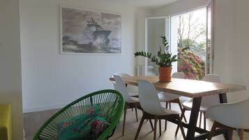 Un coin repas avec une table en bois, des chaises blanches, une chaise en osier verte, une plante en pot et un grand tableau représentant un navire accroché au mur.
