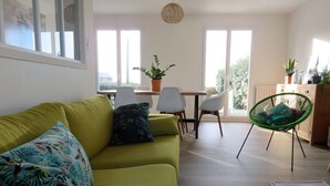 Un salón luminoso con un sofá verde, una mesa de comedor con sillas, y un sillón verde con cojín.