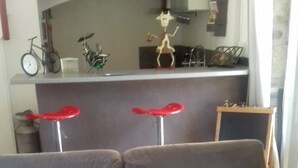 A living room with a grey sofa, a small table with a red table runner, and a kitchen area in the background. - Penty for rent in a small village typical of the peninsula (crozon)