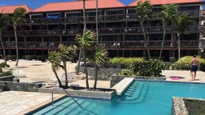 A swimming pool with stone steps and a stone deck, surrounded by palm trees and a building with balconies. - Your Perfect Beachfront Condo in Paradise - Sapphire Beach - Corner Unit (St Thomas)