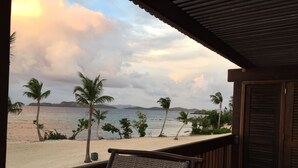 A beach view from a covered outdoor area with a wooden deck, a wicker chair, and a table with a glass. - Your Perfect Beachfront Condo in Paradise - Sapphire Beach - Corner Unit (St Thomas)